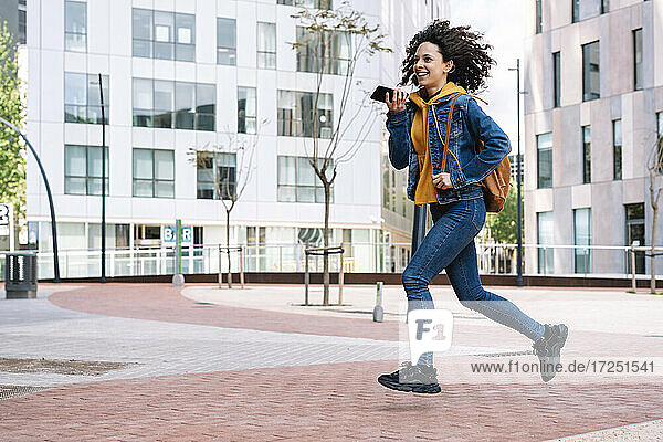 Smiling young woman with backpack talking on mobile phone while running in city