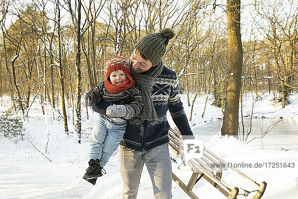 Happy father carrying son while holding sled on snow during winter