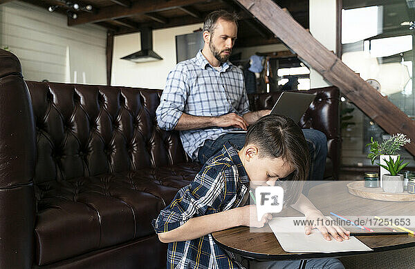 Boy drawing on paper while father using laptop on sofa at home