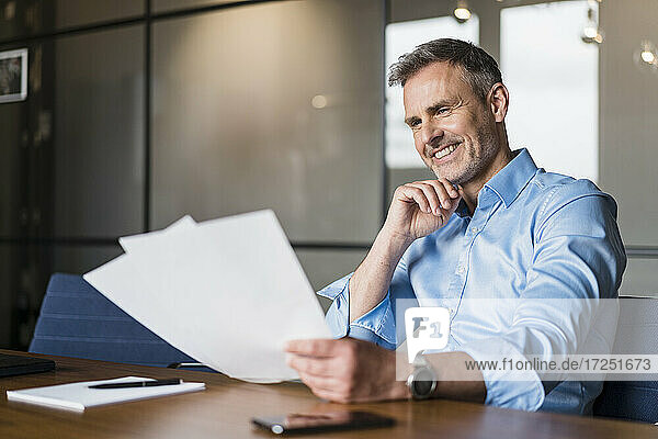 Smiling male professional with hand on chin examining paper documents in office