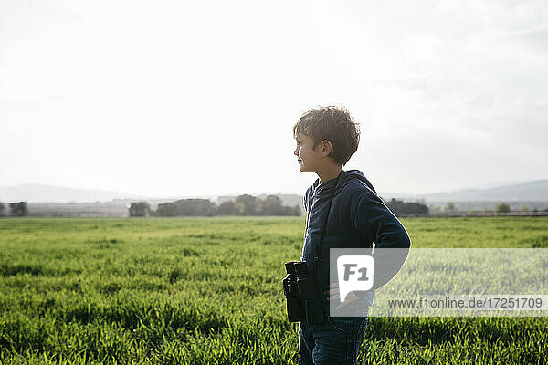 Cute boy with binoculars looking away while standing in field during sunny day