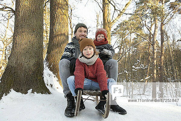 Father sitting on sled with sons near tree trunk during winter