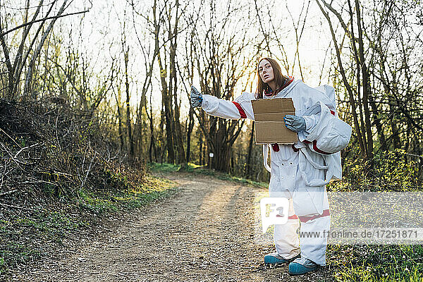 Weibliche Astronautin im Raumanzug gestikuliert  während sie einen Karton auf einem Feldweg hält