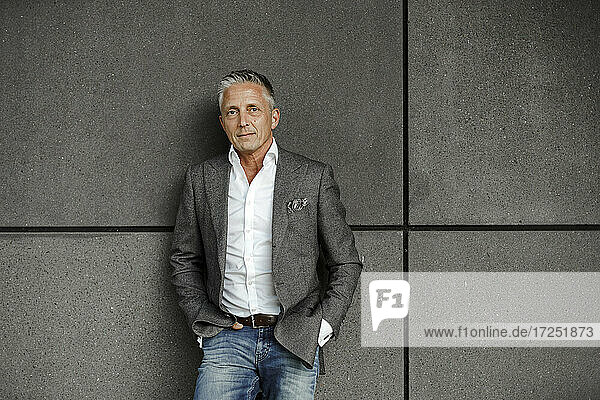 Male entrepreneur with hands in pockets leaning on gray wall