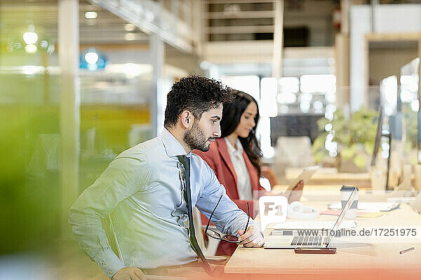 Männlicher Unternehmer  der eine Brille hält  während eine Kollegin in einem Coworking-Büro auf ihren Laptop schaut