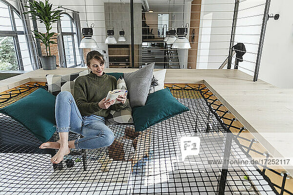 Eine Frau liest ein Buch  während sie auf einer Hängematte in einem modernen Haus sitzt