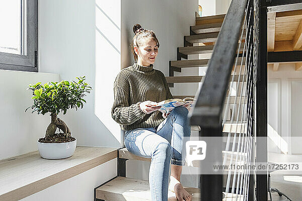 Woman reading book on staircase at home during sunny day