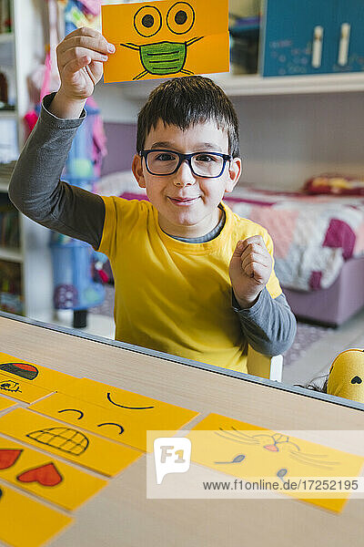 Junge mit Brille hält Emoticon über dem Kopf beim Spielen zu Hause