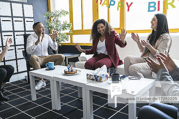 Lächelnde Geschäftsfrau  die ihren Kollegen  die während einer Geburtstagsfeier im Büro klatschen  eine Stopp-Geste zeigt