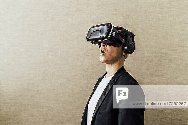 Geschäftsfrau steht mit offenem Mund  während sie ein Virtual-Reality-Headset an der Wand im Büro benutzt