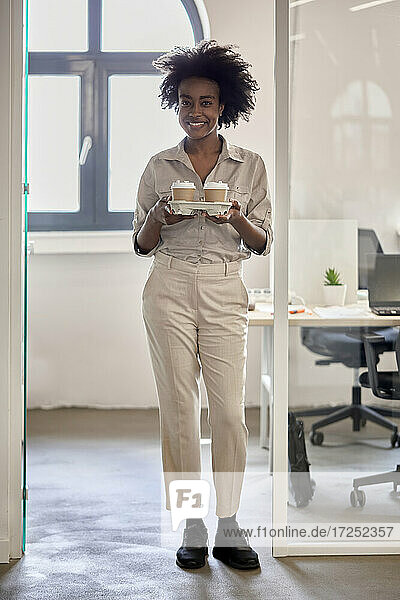 Afro businesswoman holding disposable coffee cups while standing at doorway in office