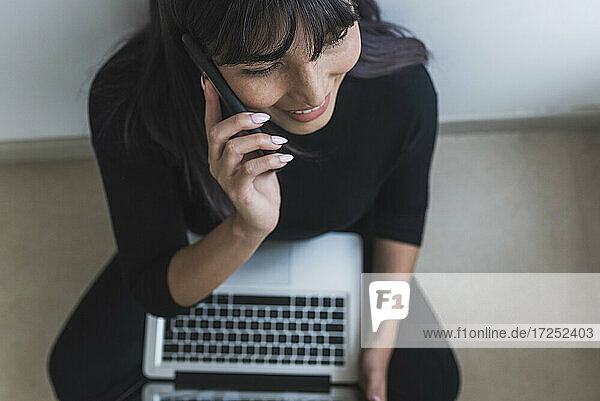 Mittlere erwachsene Frau  die mit dem Handy telefoniert  während sie zu Hause am Laptop sitzt