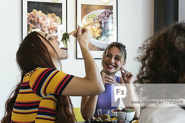 Lächelnde junge Frau sieht ihre Freundin an  die im Restaurant mit Stäbchen isst