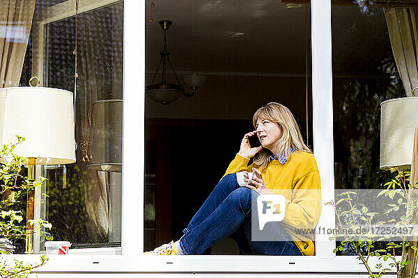 Reife Frau  die auf der Fensterbank zu Hause sitzt und mit ihrem Smartphone spricht
