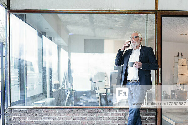 Businessman with coffee cup talking on smart phone while leaning on glass wall at office