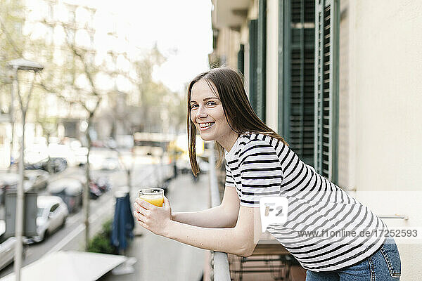Happy female tourist with glass of juice leaning on railing in balcony
