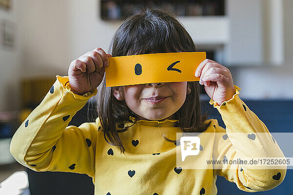 Mädchen hält gelbes Papier mit zwinkerndem Emoticon zu Hause