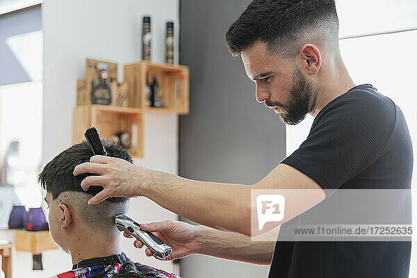 Male barber using machine to cut hair of teenage boy at salon