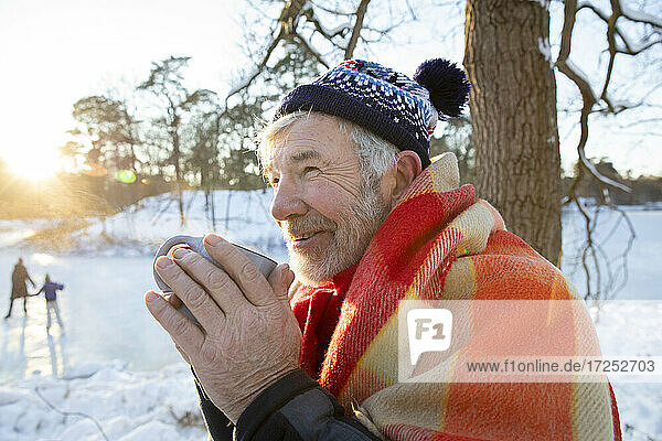 In eine Decke eingewickelter älterer Mann  der im Winter mit einer Tasse in der Hand wegschaut