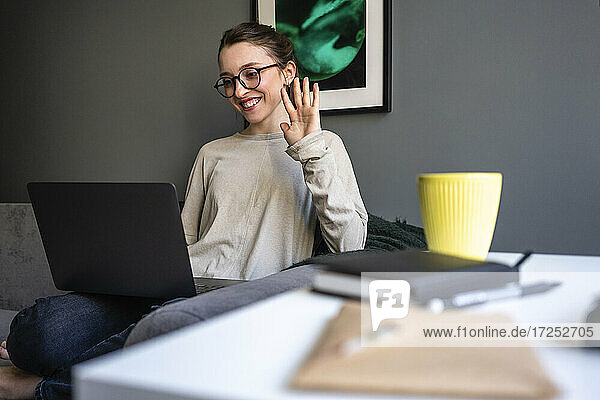 Lächelnde Frau winkt mit der Hand zum Videoanruf auf dem Laptop im Wohnzimmer