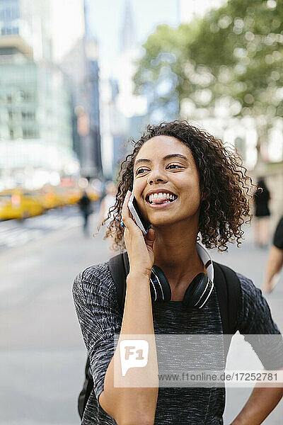 Young woman sticking out tongue while talking on smart phone in city