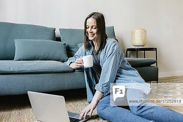 Lächelnde junge Frau  die einen Laptop benutzt  während sie zu Hause auf dem Sofa Kaffee trinkt