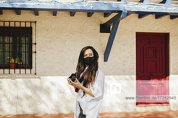 Young female tourist wearing protective face mask holding camera while standing by house at village