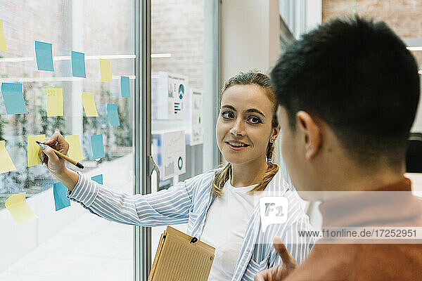 Smiling female and male entrepreneurs discussing over adhesive notes on glass wall at coworking office