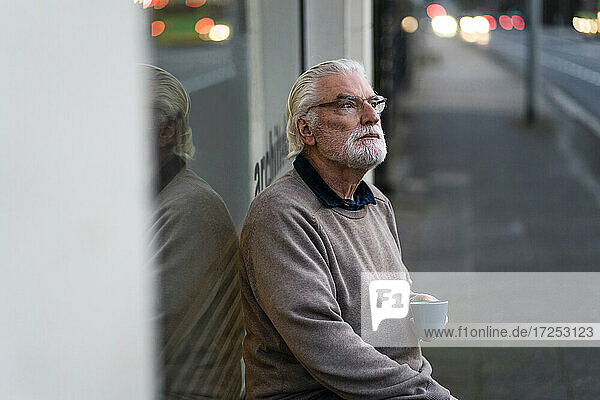 Älterer Mann mit Tasse  der wegschaut  während er außerhalb eines Cafés an der Straße sitzt