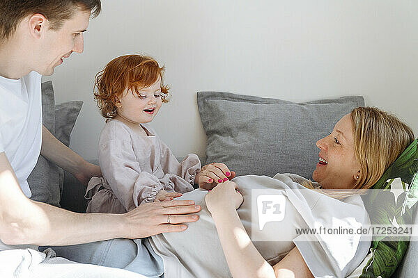 Lächelnder Vater und Tochter berühren den Bauch der schwangeren Frau auf dem Sofa zu Hause