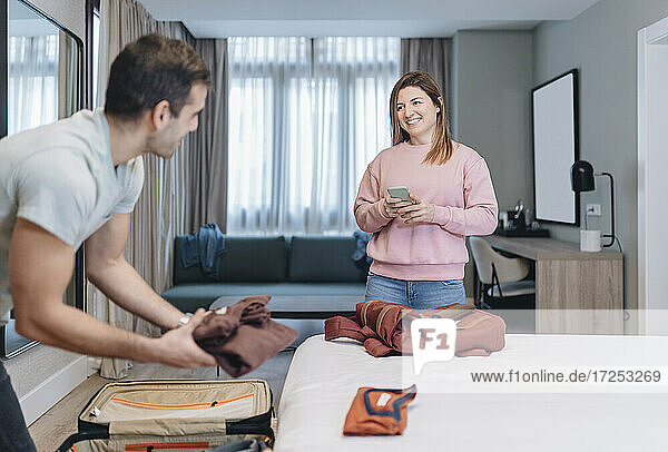 Smiling woman looking at man unpacking luggage in hotel room