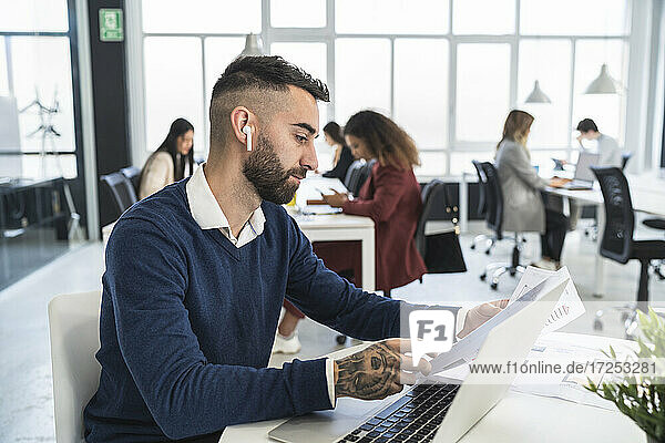 Ein männlicher Unternehmer liest ein Dokument  während er vor einem Laptop sitzt und mit männlichen und weiblichen Kollegen in einem Coworking-Büro arbeitet.