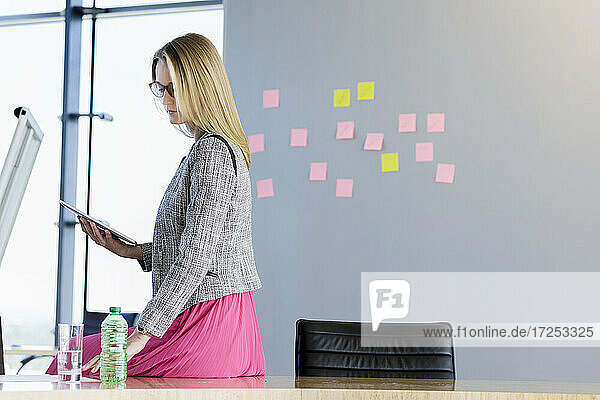 Geschäftsfrau sitzt auf einem Tisch und hält ein digitales Tablet im Sitzungssaal eines Büros