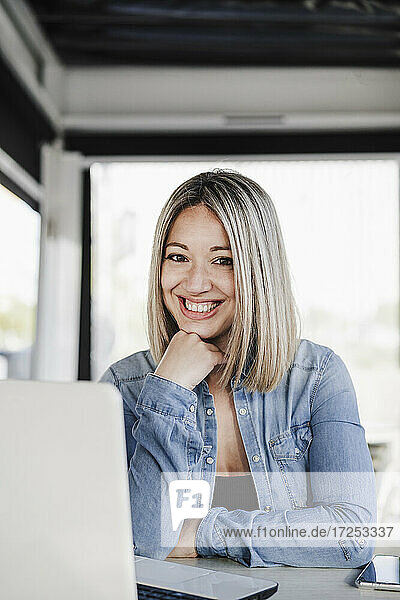 Happy businesswoman with hand on chin sitting with laptop at coffee shop