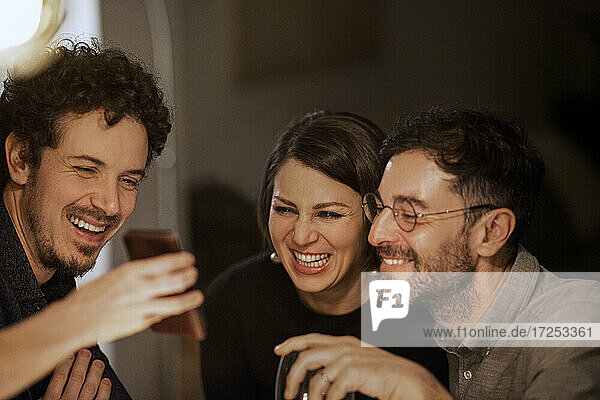 Ein glücklicher Mann zeigt seinen Freundinnen und Freunden zu Hause sein Mobiltelefon
