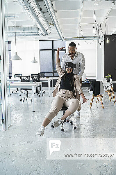 Afrikanischer Geschäftsmann steht hinter einer lächelnden Kollegin  die einen Virtual-Reality-Simulator in einem Coworking-Büro trägt