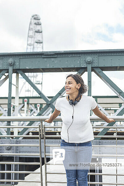Happy woman with headphones standing in front of railing on bridge  London  England  UK