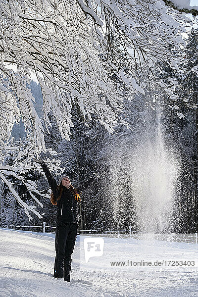 Junge Frau mit erhobenen Armen unter einem Baum auf verschneitem Land im Winter stehend