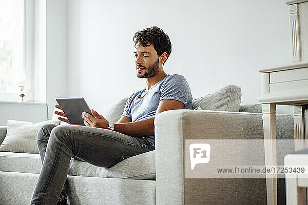 Junger Mann benutzt ein digitales Tablet  während er auf dem Sofa im Wohnzimmer sitzt