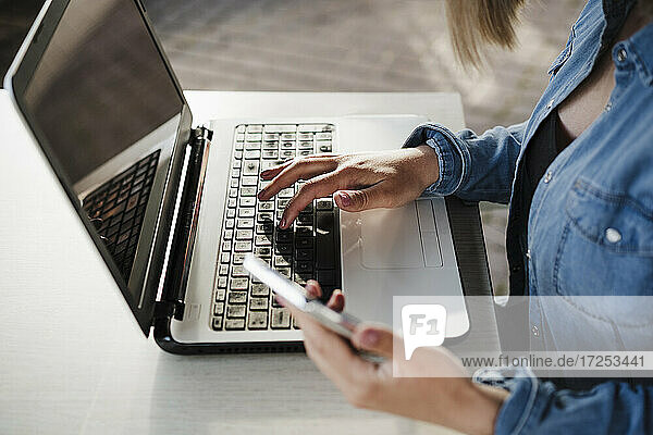 Female entrepreneur holding mobile phone while working on laptop at coffee shop
