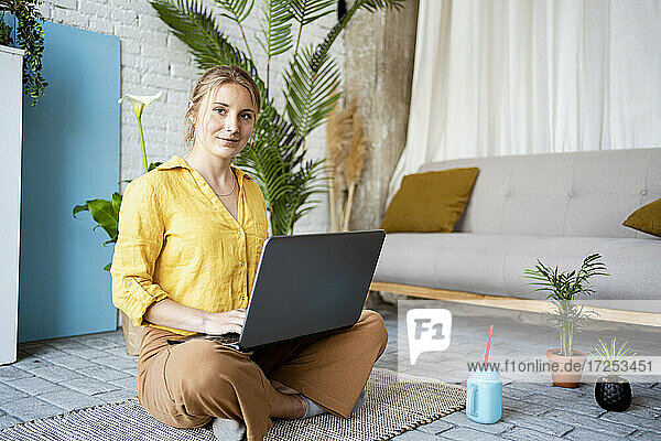 Berufstätige Frau arbeitet am Laptop  während sie zu Hause auf einer Matte sitzt