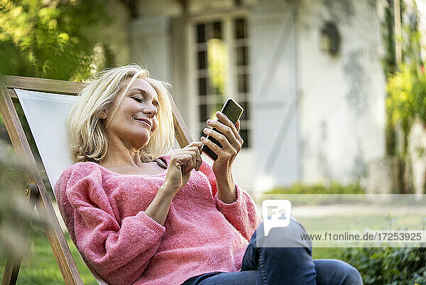 Lächelnde reife Frau  die ein Smartphone benutzt  während sie auf einem Liegestuhl sitzt
