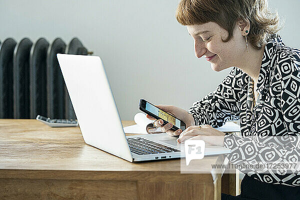 Lächelnde Frau mit Smartphone und Laptop bei der Arbeit von zu Hause aus