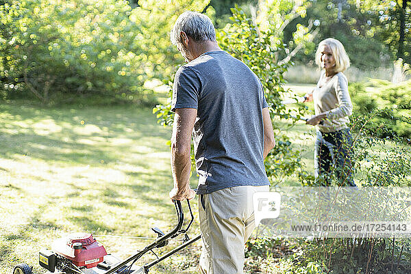 Älterer Mann mäht Rasen im Garten  während seine Frau im Hintergrund arbeitet