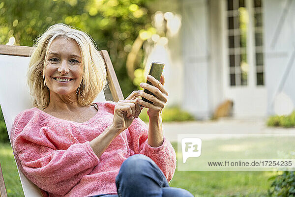 Porträt einer lächelnden reifen Frau  die ein Smartphone benutzt  während sie auf einem Liegestuhl sitzt