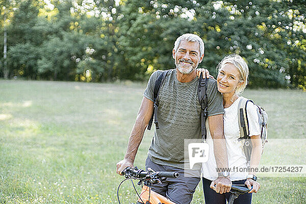Porträt eines lächelnden reifen Paares mit Fahrrad im Wald stehend