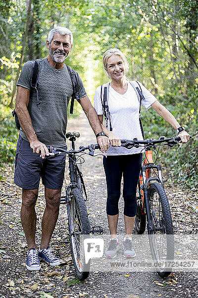 Portrait of smiling mature couple with bicycles standing on footpath in forest