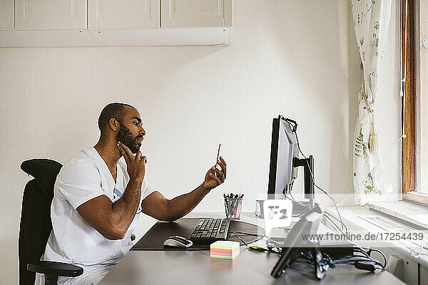 Männlicher Arzt  der einen Videoanruf über ein Smartphone tätigt  während er am Schreibtisch sitzt
