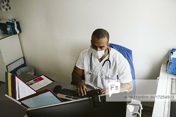 Männlicher Mitarbeiter im Gesundheitswesen mit Gesichtsmaske bei der Arbeit am Computer in einer medizinischen Klinik