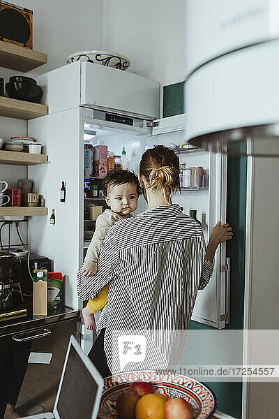 Rückansicht einer Mutter  die einen kleinen Jungen trägt  während sie den Kühlschrank in der Küche öffnet